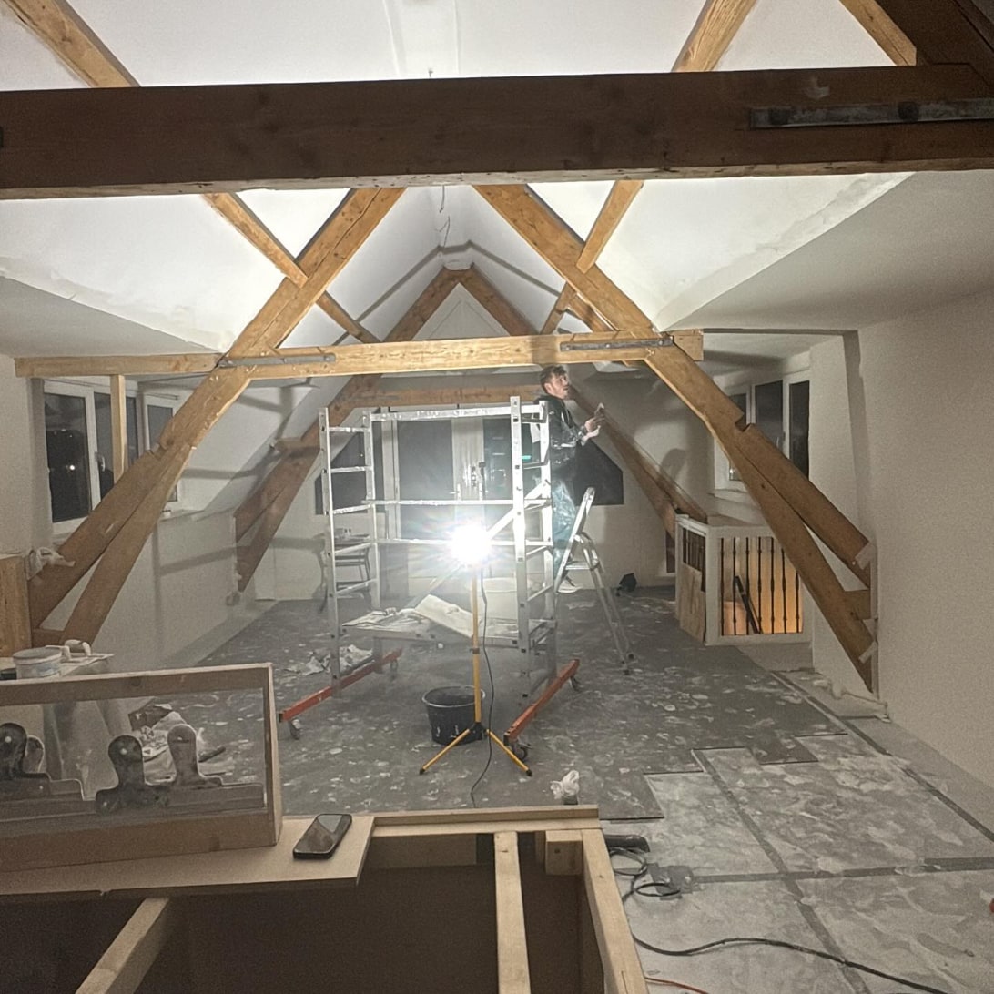Zolder met oude houten spanten en nieuw wit stucwerk tussen de balken, met een rolsteiger in het midden